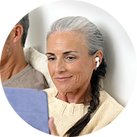 A woman with gray hair, wearing an in-ear hearing aid, sits indoors next to a man while holding a tablet. She wears a beige sweater, and the setting appears relaxed and homey.