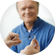 A person holds a hearing aid, presenting it to an older man who smiles and looks on, in a brightly lit room.