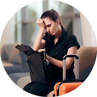 A woman sits in an airport lounge, resting her head on her hand while searching through a bag; an orange suitcase is placed beside her.
