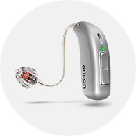Silver hearing aid labeled "Oticon" with an earpiece, connected by a thin wire, positioned on a light gray circular background. The device has a small green indicator light.