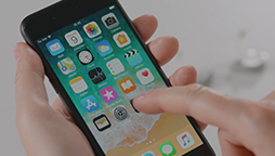 A hand holds a smartphone, navigating through various application icons on the home screen, in an indoor setting.