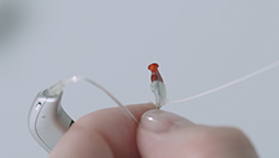 Small hearing aid with a clear plastic tube and a red-tipped earpiece held between fingers against a neutral light grey background.