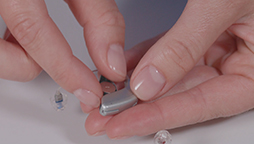 Hands holding a small, silver hearing aid while adjusting it on a white surface, with additional hearing aid components nearby.