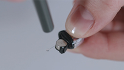 A hand holds a tiny hearing aid device close to a small black tool, possibly during a repair or adjustment, in a well-lit environment.