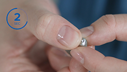 Hands gently holding a small hearing aid with a blue circle displaying '2 SEC' in the upper left corner. The background features blurry blue clothing.