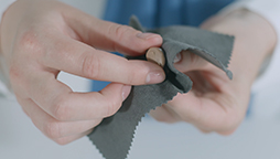 Hands using a gray microfiber cloth to clean a small coin, with a blurred background of blue and white attire.