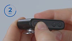 Hands pressing buttons on a small black hearing device, highlighted by a blue '2 SEC' indicator on a gray background.