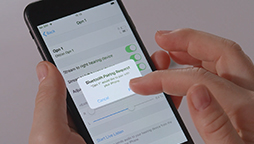 Hands interact with a black smartphone displaying settings. A pop-up message reads, “Bluetooth Pairing Request: ‘APP-Device’ would like to pair with your iPhone.” The background is plain and light-colored.