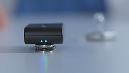 A black hearing device with two glowing blue lights sits on a white surface, with a metallic hearing aid blurred in the background.
