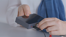 Hands connecting a cable to a compact electronic device, with a blue fabric and white sleeve in the background.
