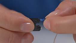 Hand inserting a blue component into a small black hearing device, close-up against a blue background.