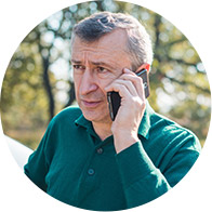 A man in a green shirt talks on a smartphone outside, with trees and sunlight in the background.