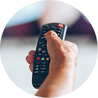 A hand holds a TV remote, pressing a button; the background shows a blurred television screen, creating a relaxed, home entertainment context.