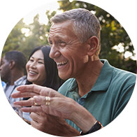 A smiling elderly man in a green polo shirt gestures animatedly while sitting outdoors among people, with trees and sunlight in the background.