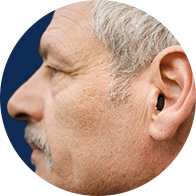 A close-up of an elderly man's left side profile, featuring a small hearing aid in his ear against a solid dark blue background.