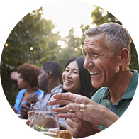 People are smiling and conversing, seated around an outdoor table, against a backdrop of greenery with blurred lights.