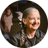 An older woman with gray hair and a braid smiles while talking to another person in a dimly lit indoor setting. Both wear dark clothing.