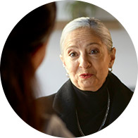 An older woman with white hair and a black outfit speaks to another person in a well-lit, indoor setting with blurred background elements.