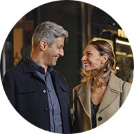 A gray-haired man and a woman in professional attire are walking together, possibly outdoors at night, amidst urban lighting. The image promotes hearing care services, with no visible branding or text.