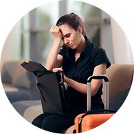A woman sits in an airport lounge, resting her head on her hand while searching through a bag; an orange suitcase is placed beside her.