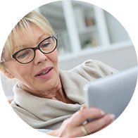 A woman wearing glasses and a scarf is reading a tablet device while sitting indoors in a bright, white room with shelves in the background.