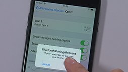A hand operates a smartphone showing a "Bluetooth Pairing Request" pop-up, asking to connect to "Opm 1" hearing device. Several toggles are visible in the phone's hearing device settings.