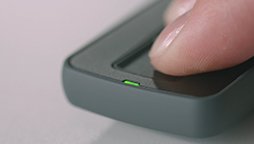 A finger presses against a fingerprint scanner on a compact black device, illuminated by a small green light, positioned on a grey surface.