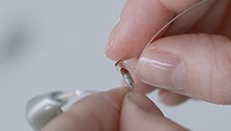 Hands hold a small hearing aid, adjusting a tiny wire in a minimal, possibly clinical environment.
