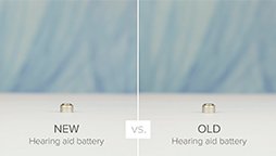 Two small, cylindrical hearing aid batteries are positioned side by side on a white surface against a blurred blue background. Text: "NEW Hearing aid battery" on the left and "OLD Hearing aid battery" on the right, with "vs." in the center.