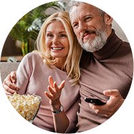 Couple sitting on a sofa sharing a bowl of popcorn and holding a remote control.