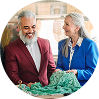 Two individuals with gray hair, wearing red and blue blazers, examine a green patterned fabric in a well-lit room filled with textiles and creative supplies, suggesting an artisan or boutique setting.