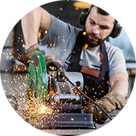 A man wearing ear protection and gloves cuts metal with an angle grinder, creating sparks, in a workshop filled with tools and equipment in the background.