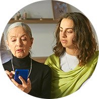 An older woman uses a smartphone while a younger woman observes beside her, both indoors with shelves in the background.