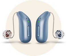 Two blue hearing aids, each with a small, clear speaker attached by a thin wire, sit upright on a light beige circular background. Green indicator lights are illuminated on both devices.