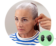 A person with gray hair is being fitted with a hearing aid by an audiologist. The context suggests a professional hearing clinic. A small image of hearing aids is displayed in the corner. No visible branding or text is present.
