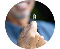 A hand holds a discreet hearing aid between fingers, highlighting its small size. In the background, a smiling man blurs into focus, signaling satisfaction and advanced hearing care solutions.