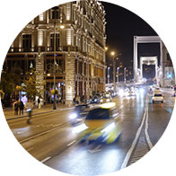 Vehicles travel down a busy, illuminated street at night, passing by a grand, ornate building on the left with pedestrians nearby and a large bridge in the background.