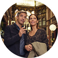 A couple stands closely, one holding a small device resembling a hearing aid, in a well-lit, ornate indoor setting featuring decorative archways, warm lighting, and vintage-style elements. No visible branding or text.