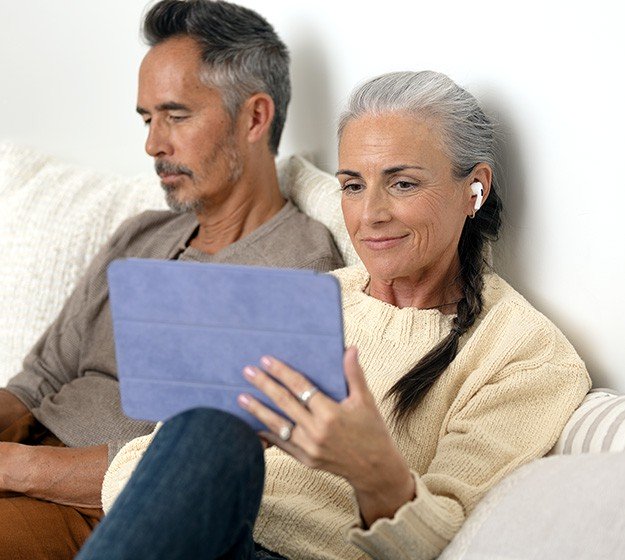 A woman wearing a hearing aid sits on a couch beside a man, holding a tablet with a blue cover. They appear engaged in a relaxed home environment.