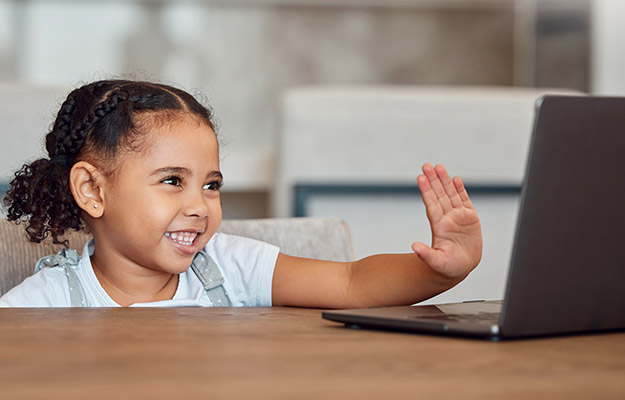 A smiling child waves excitedly at a laptop while sitting at a wooden table in a brightly lit, modern indoor space, suggesting communication, virtual interaction, or a remote session.