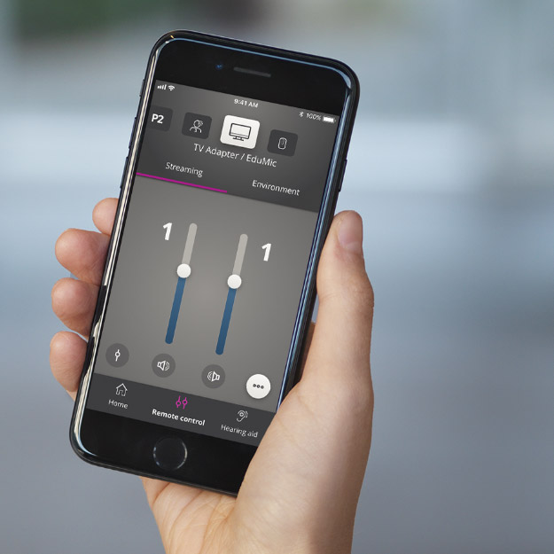 Smartphone screen displays a hearing aid remote control app with adjustable volume sliders, streaming options, and icons for TV Adapter/EduMic against a blurred indoor background.