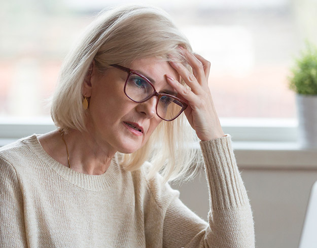 Woman with blonde hair wearing glasses, holding her forehead, appearing to experience discomfort or pain.