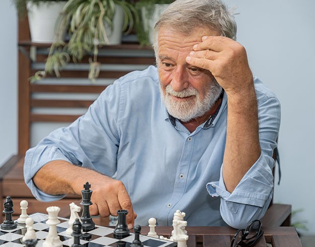 An older man focuses intently on a chess game, seated on an outdoor patio surrounded by greenery.