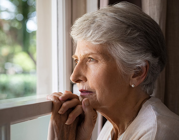 Older woman with short gray hair gazing out of a window, hands resting on the windowsill.