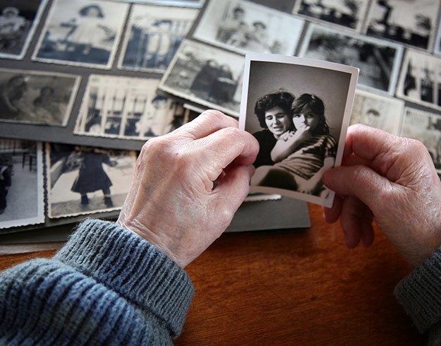 Elderly hands holding a black-and-white photo in front of a table covered with vintage family pictures.