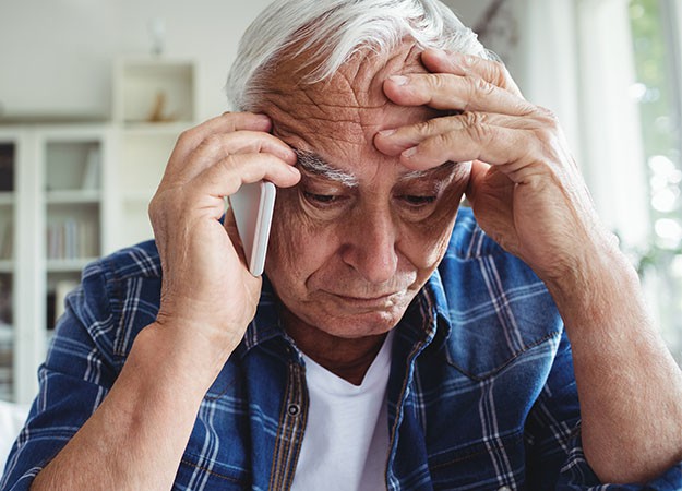 Older man holding his head with one hand while speaking on a phone, showing signs of distress or discomfort.