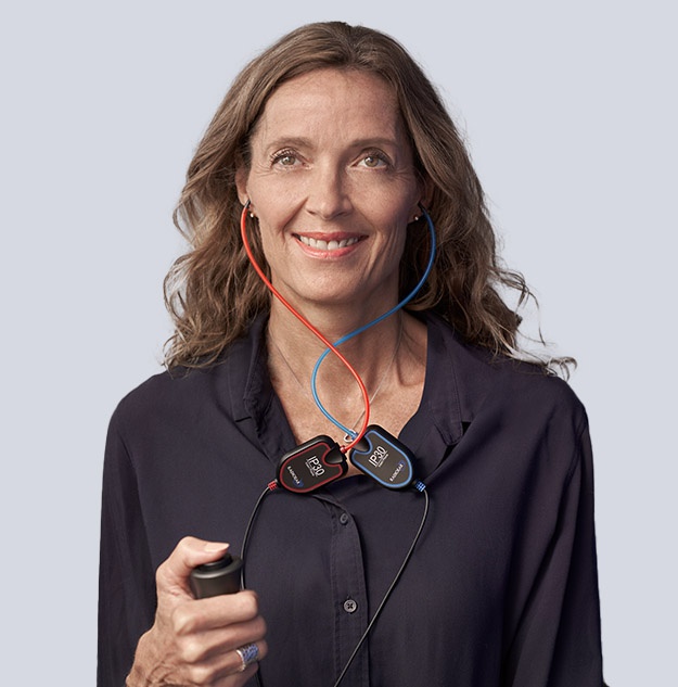 A smiling woman uses a handheld hearing care device with attached red and blue labeled wires.