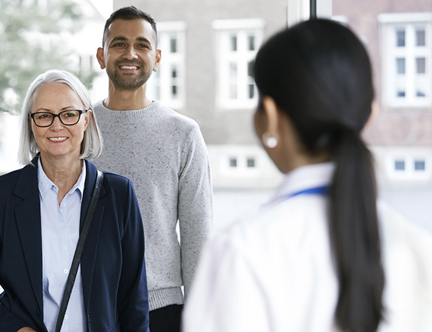 Two adults, an older woman and a younger man, smiling in a bright indoor setting while engaging with a healthcare professional wearing a white coat.