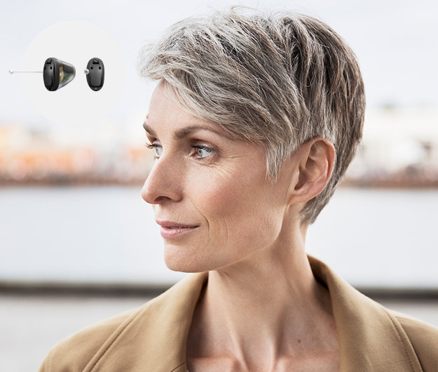 Middle-aged woman with short grey hair gazing to her left in an outdoor urban setting, with small hearing aids displayed in the top-left corner.
