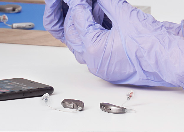 Hearing aids on a table near a smartphone, with purple gloved hands adjusting another hearing aid in the background.